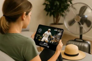 Woman watching live Wimbledon tennis match on IPTV using a tablet, sitting in a cool summer setting with fan, suitcase, and sunhat nearby